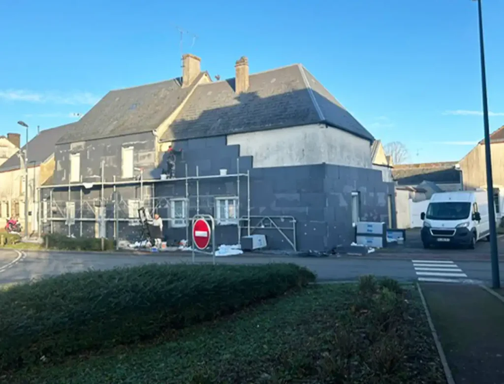 Une maison en cours de rénovation sur un chantier d'isolation par l'extérieur, avec des échafaudages et des panneaux d'isolation sur les murs extérieurs. Des matériaux de construction sont éparpillés sur le sol, une camionnette blanche est garée à proximité et un panneau d'interdiction d'accès se trouve devant sous un ciel dégagé.