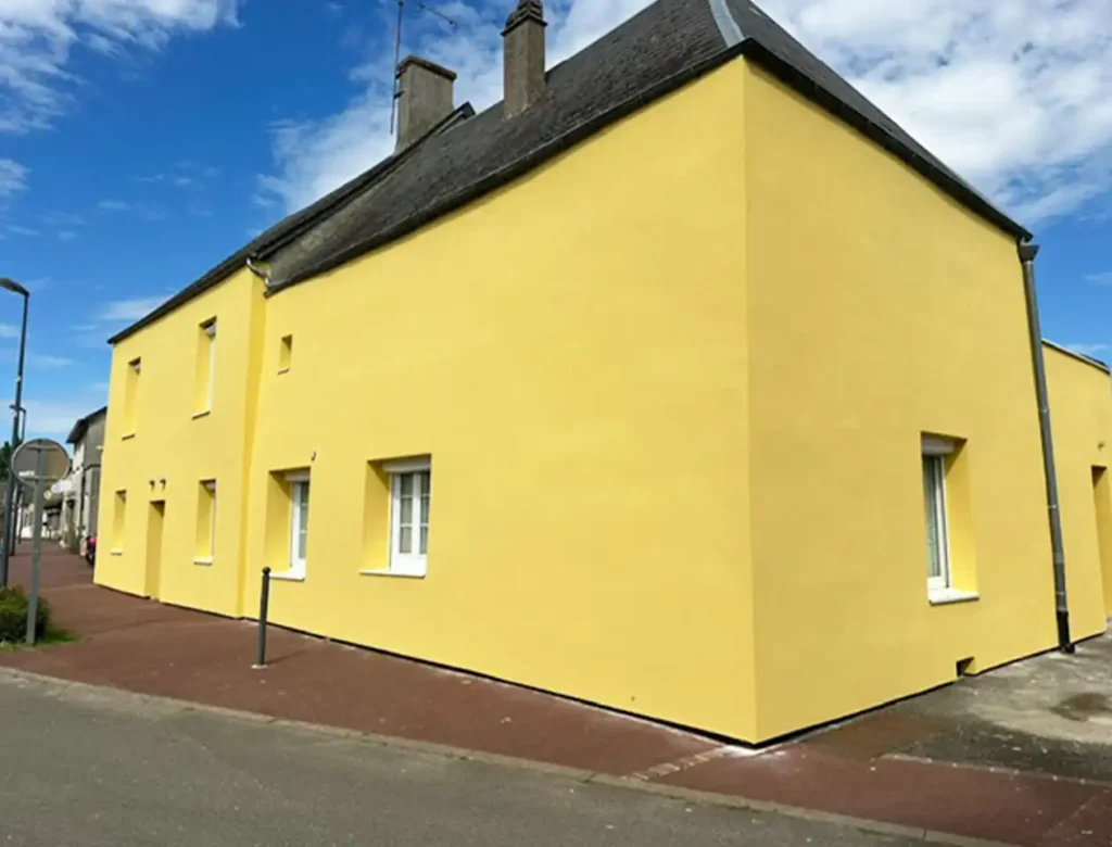 Une maison jaune vif avec des fenêtres à cadre blanc se trouve au coin d'une rue sous un ciel partiellement nuageux. Avec une fin de chantier isolée par l'extérieur, le bâtiment simple et moderne présente des murs lisses et un toit gris foncé.
