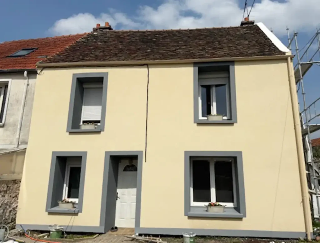 Une petite maison beige de deux étages avec un toit en tuiles brunes, des cadres de fenêtres gris et une porte blanche. Malgré la fin de chantier d'un ravalement, une fissure visible court encore au centre de la façade. Quatre fenêtres sont ornées de charmantes jardinières.
