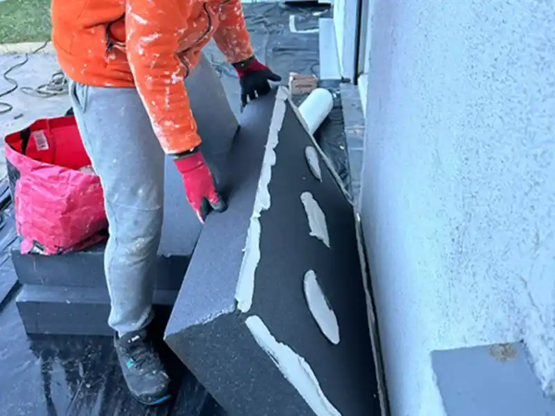 Une personne portant une veste orange et un pantalon gris manipule l'application de panneau isolant, pla&ccedil;ant un panneau isolant avec de l'adh&eacute;sif sur un mur ext&eacute;rieur. Un sac rouge et d'autres mat&eacute;riaux de construction se trouvent &agrave; proximit&eacute;.