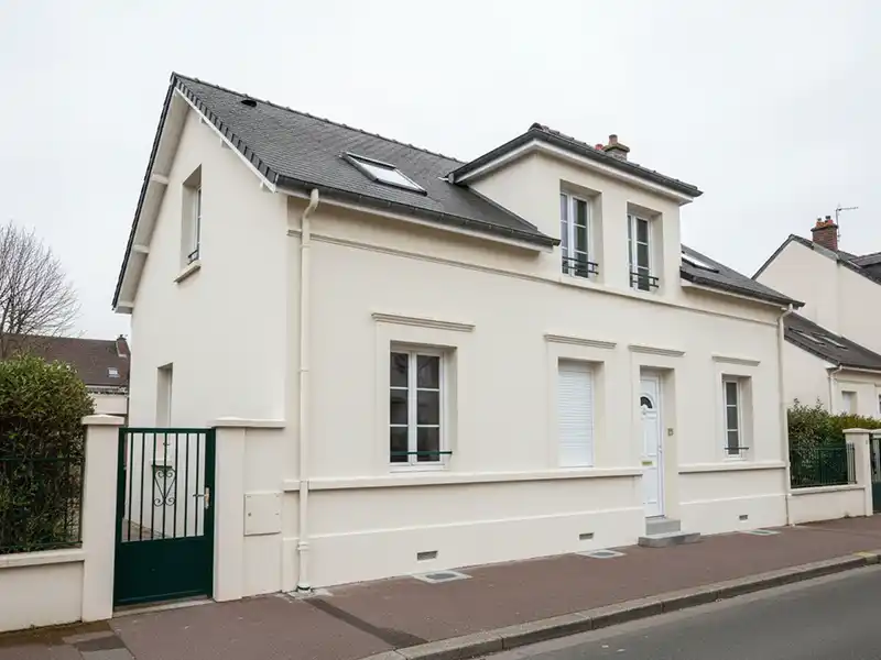 Maison repeinte - Isolex Maison à deux étages de couleur crème, fraîchement repeinte, avec un toit gris foncé et plusieurs fenêtres à cadre blanc, située dans une rue calme avec un portail vert et un trottoir devant.